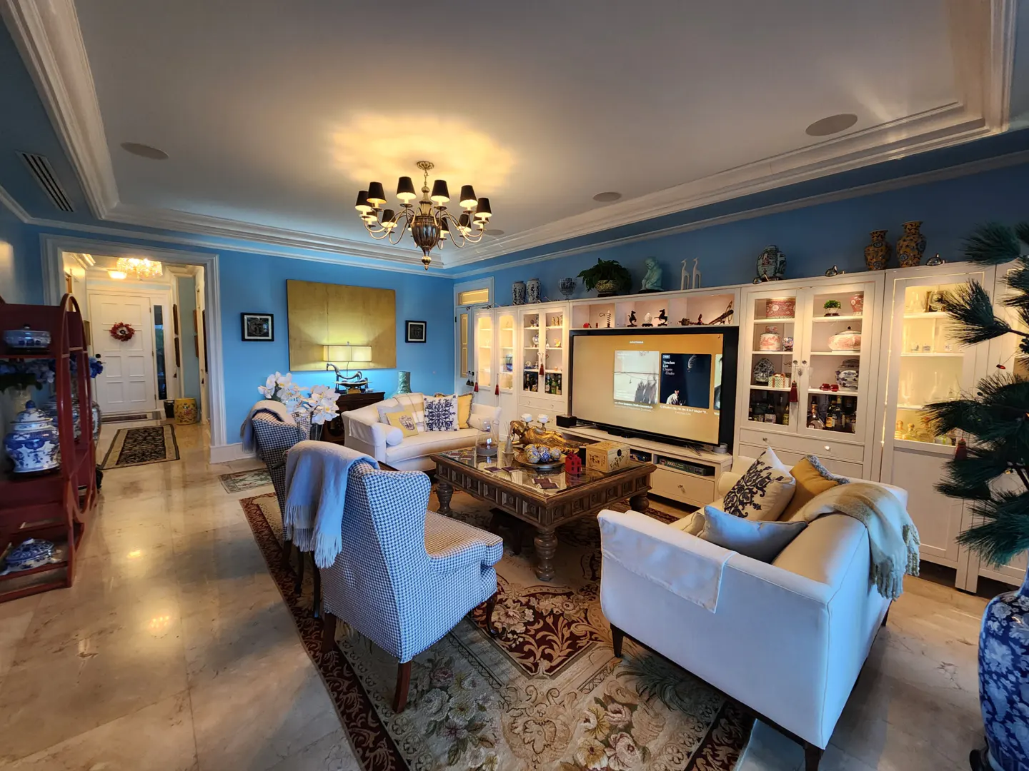 A bright living room with blue walls, white trim, and marble floors. A large TV is mounted above white cabinets. A chandelier hangs above a floral rug.