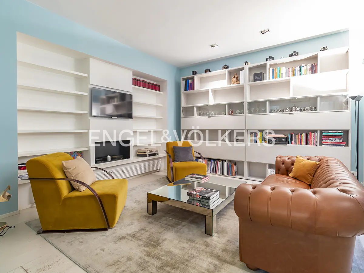 Living room with a brown leather sofa, yellow armchairs, and a glass coffee table on a gray rug. White bookshelves line the walls.