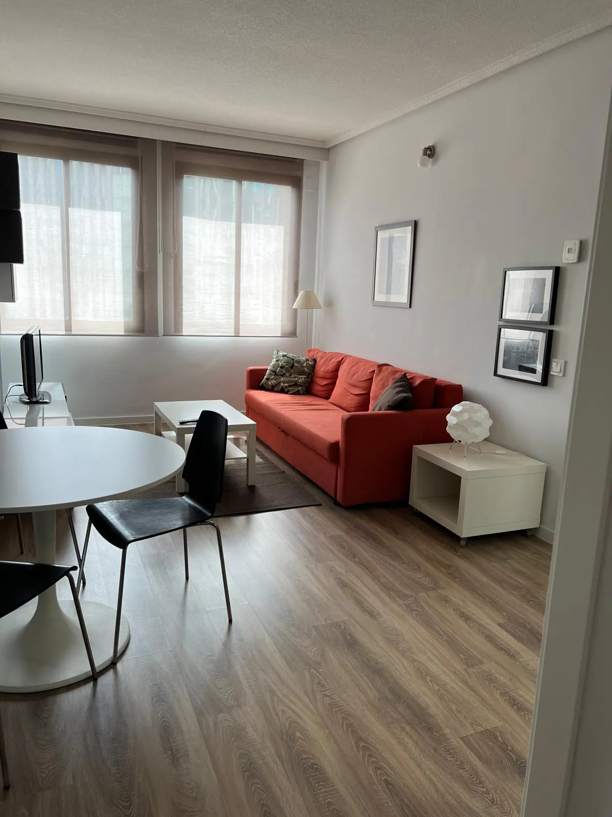 A bright living room with wood floors, an orange sofa, a white table, and black chairs. Natural light streams through the windows.