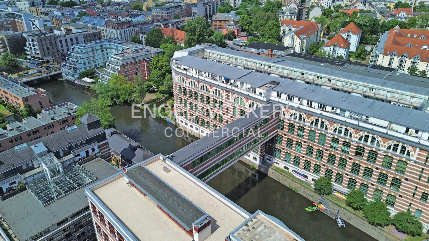 Aerial view of a large brick building connected by a glass bridge over a canal in a dense urban neighborhood.