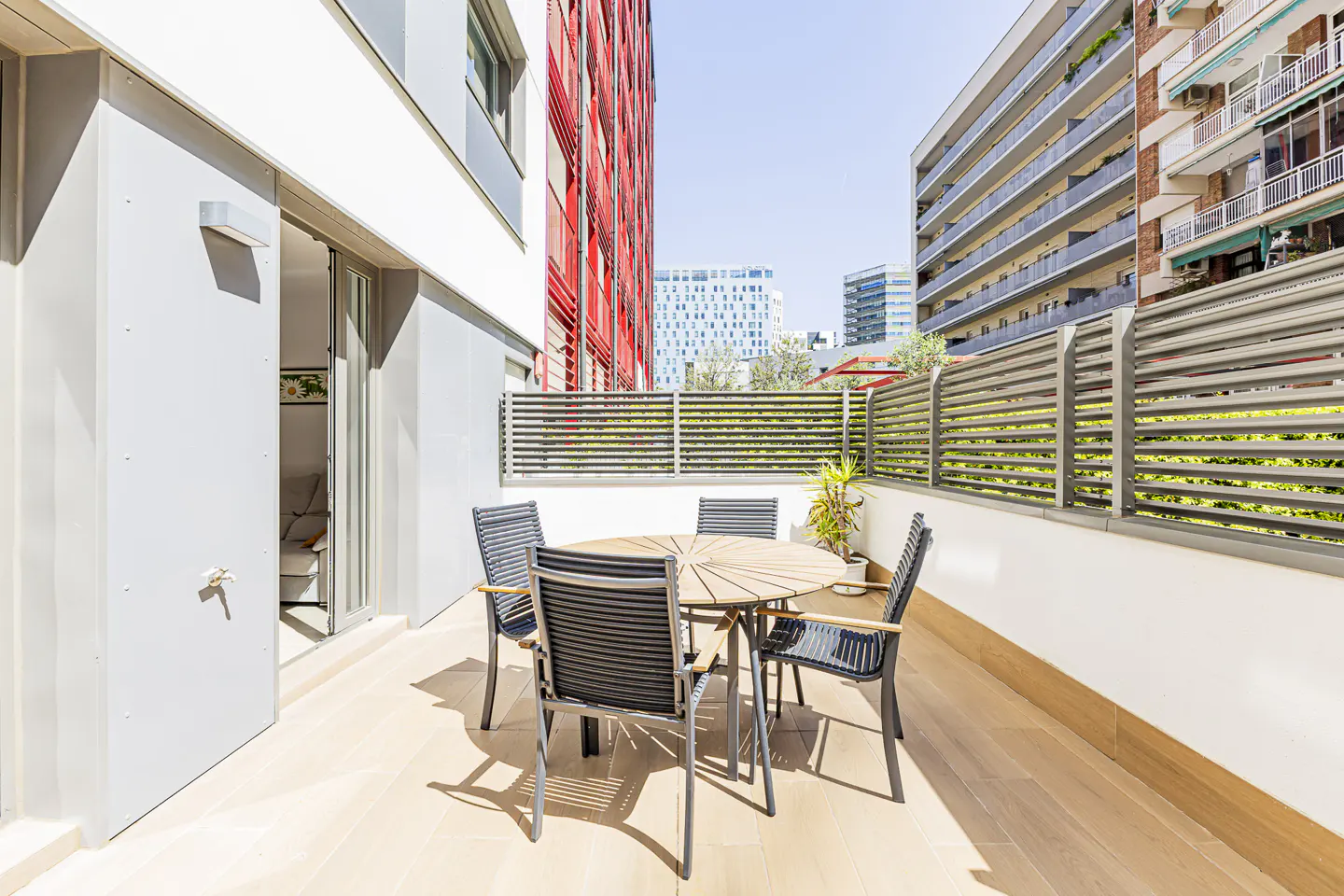 Balcony with a round wooden table and four chairs. A potted plant sits near the railing, with city buildings in the background.