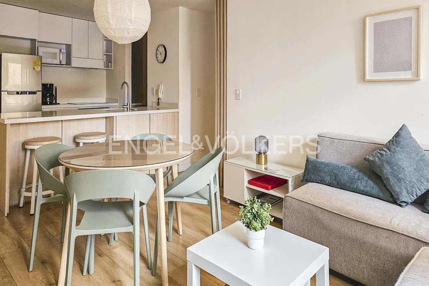 A modern apartment interior with a kitchen, dining area with green chairs, and a living room with a gray sofa and pillows.