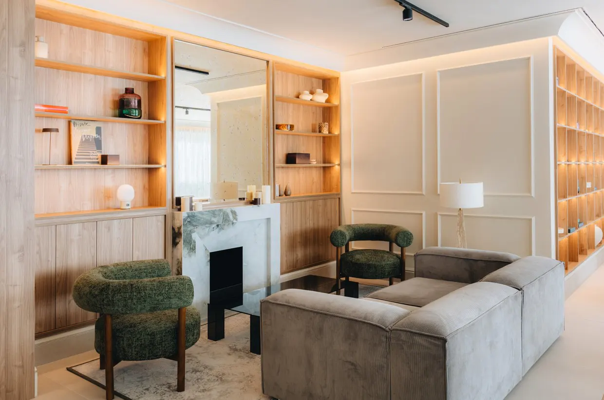 Living room with a gray sofa, green chairs, and a marble fireplace. Wooden shelves are built into the wall.