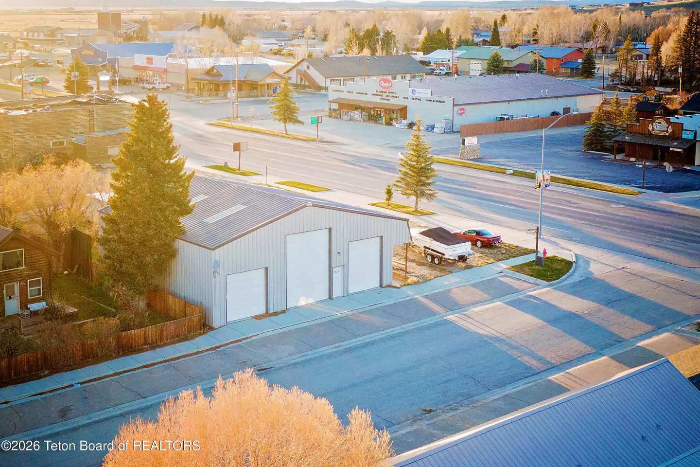 Aerial view of a metal building with three white garage doors, a trailer, and a red car, in a small town.