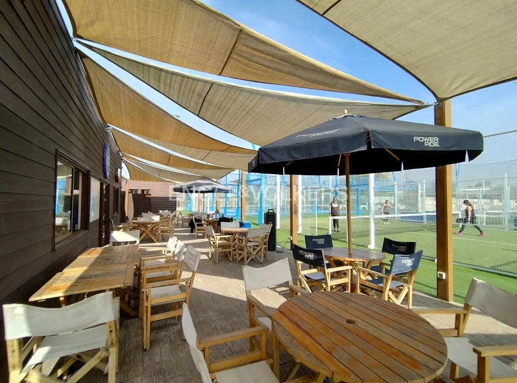 Outdoor patio with wooden tables and chairs under a beige awning. Paddle tennis court with players visible in the background.