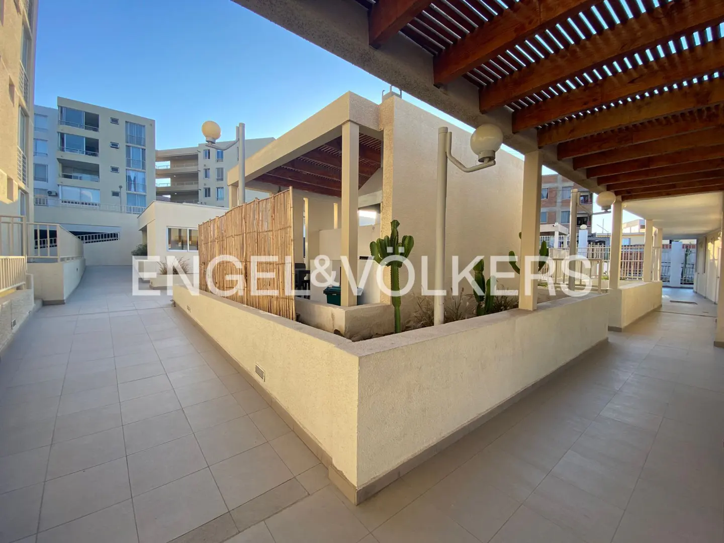 Exterior view of a modern apartment complex with tiled walkways and wooden pergolas. Cacti grow in raised planters. The Engel & Völkers logo is superimposed.