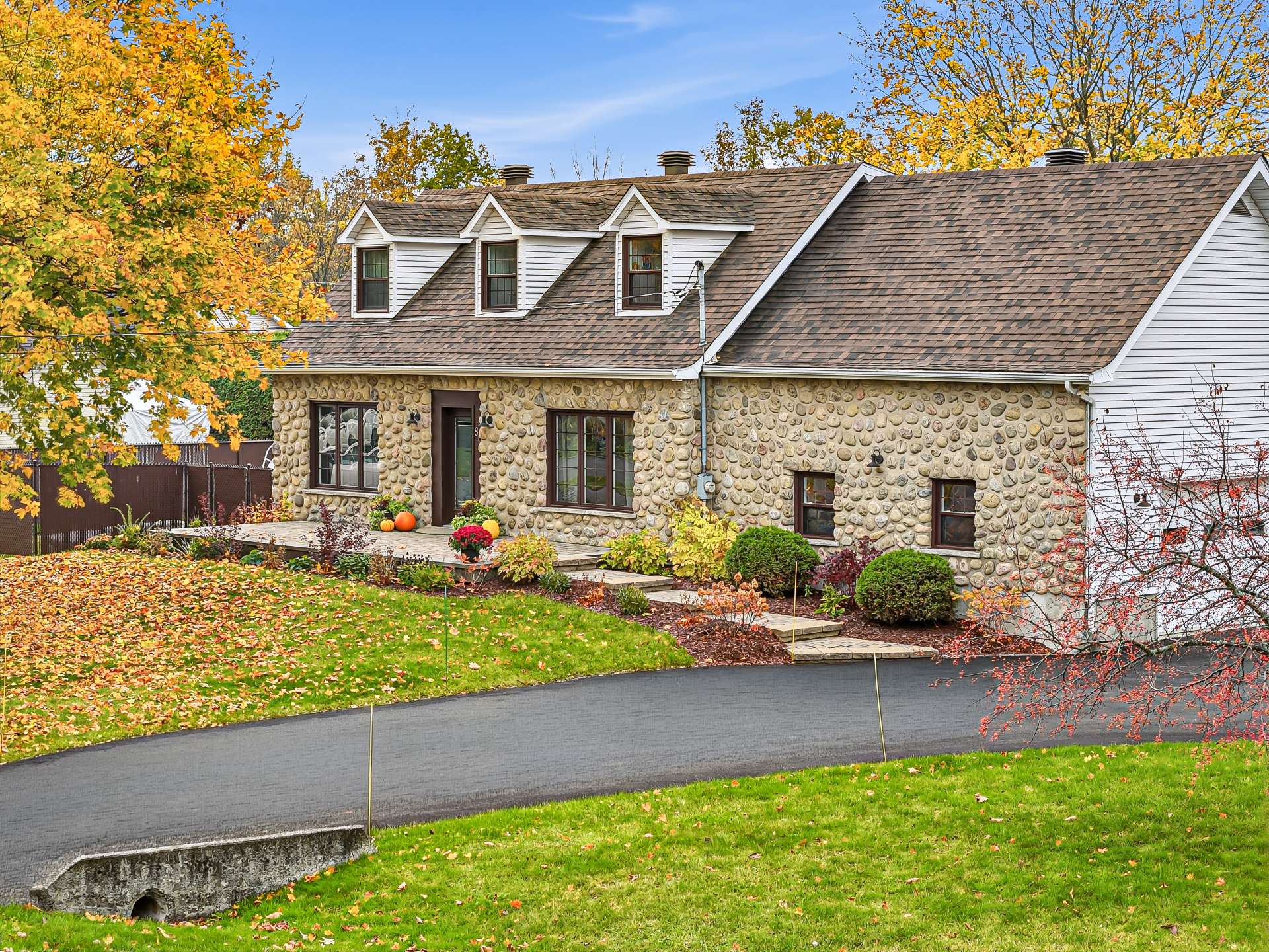 A stone-faced house with three dormers, a brown roof, and a curved driveway, surrounded by autumn trees and fallen leaves. [AI-generated text]