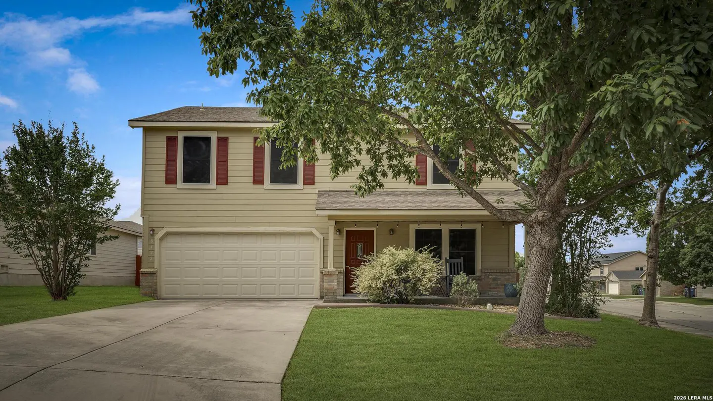 Two-story beige house with red shutters, a two-car garage, and a green lawn under a blue sky.
