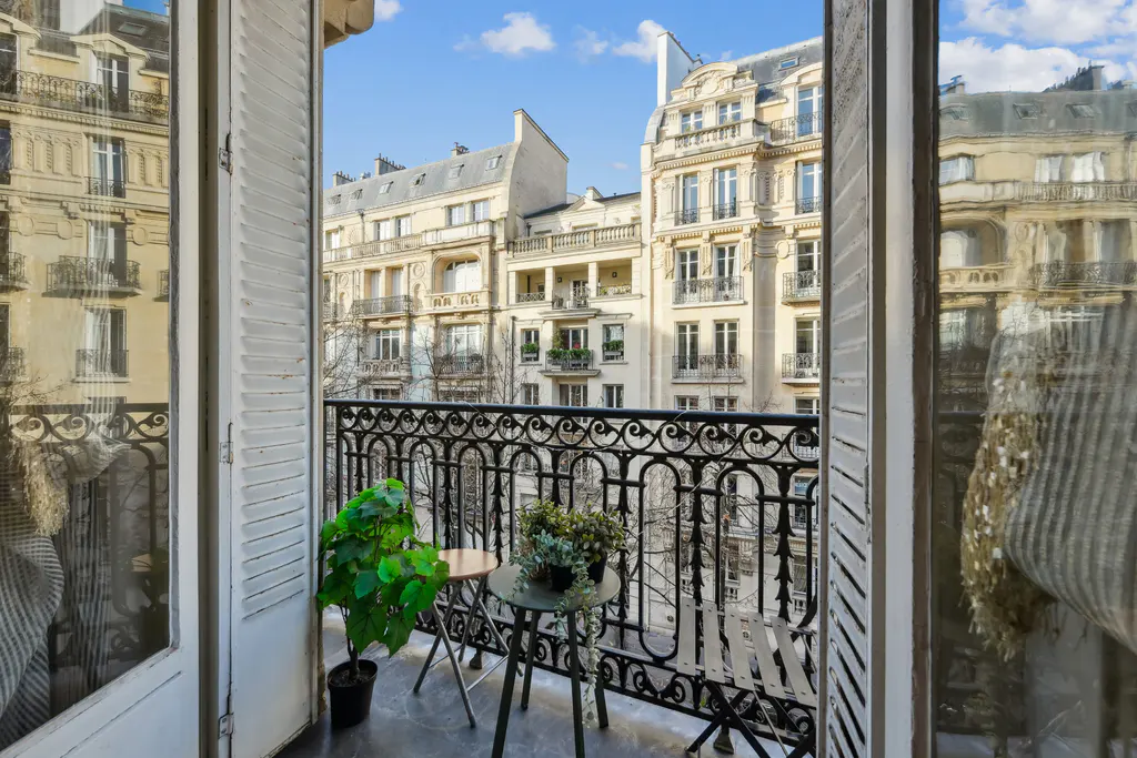 View from a Parisian balcony with black iron railing, plants, and a small table and chair set. Buildings are visible in the background.