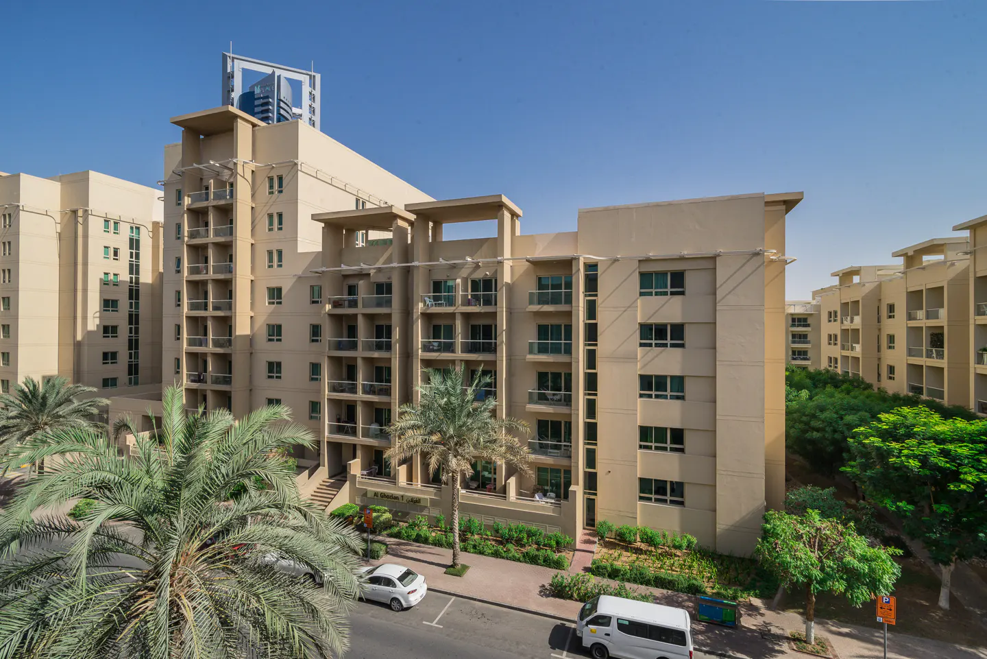 Exterior view of beige apartment buildings with balconies, palm trees, and cars parked on the street. Blue sky in the background.