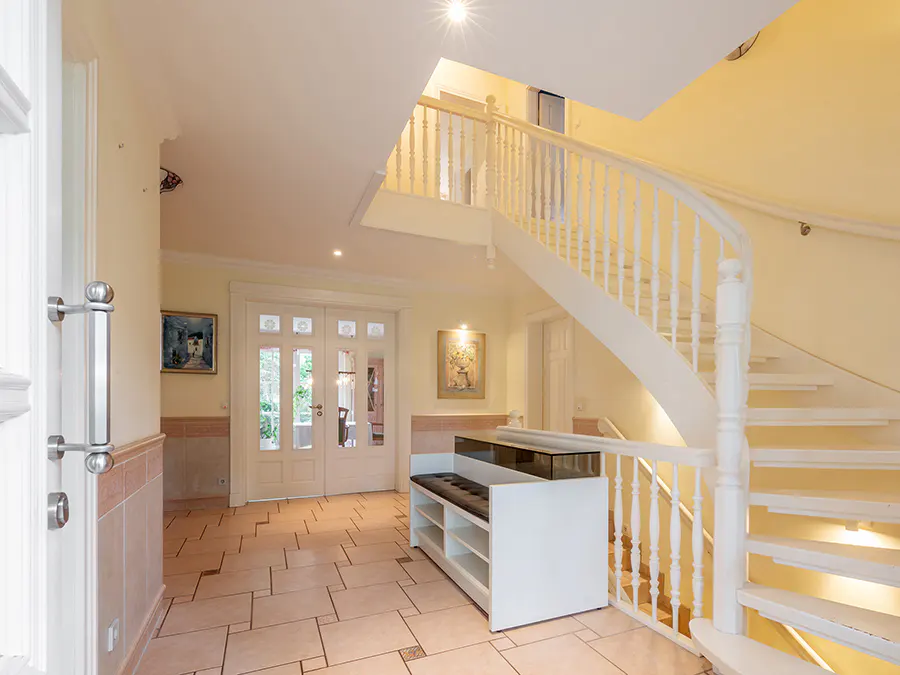 Bright foyer with white staircase, balustrade, and shoe bench. Double doors lead outside. Beige tile floor and yellow walls.