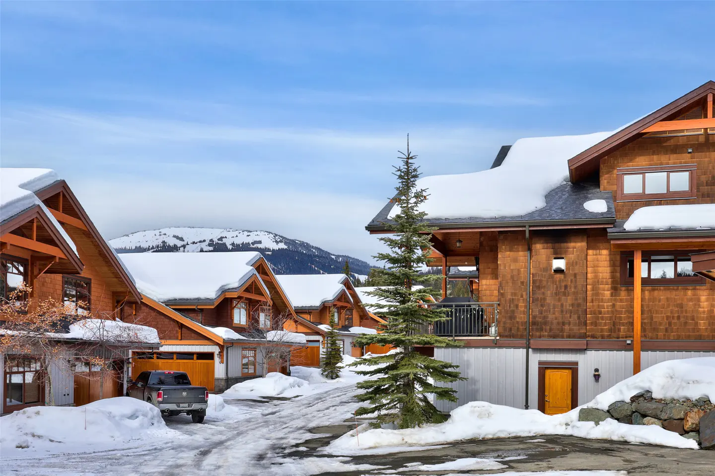 Row of brown, wood-shingled townhouses with snow-covered roofs on a winter day. A gray truck is parked in front of a garage.