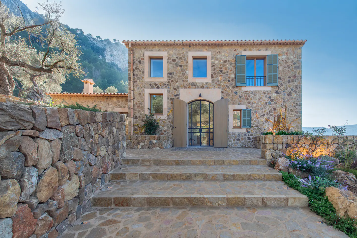 Stone house with blue shutters and a black-framed glass door. Stone steps lead to the entrance. A stone wall and trees are on the left.