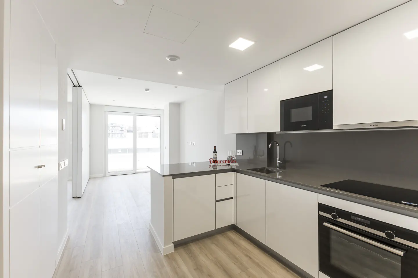 Modern kitchen with white glossy cabinets, grey countertops, built-in appliances, and an open view to a bright living area.