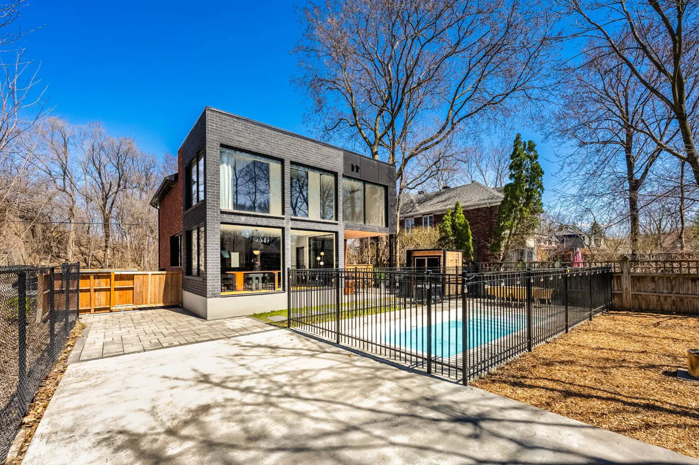 Modern two-story home with a pool. The house has large windows and a gray brick exterior. A black fence surrounds the pool.