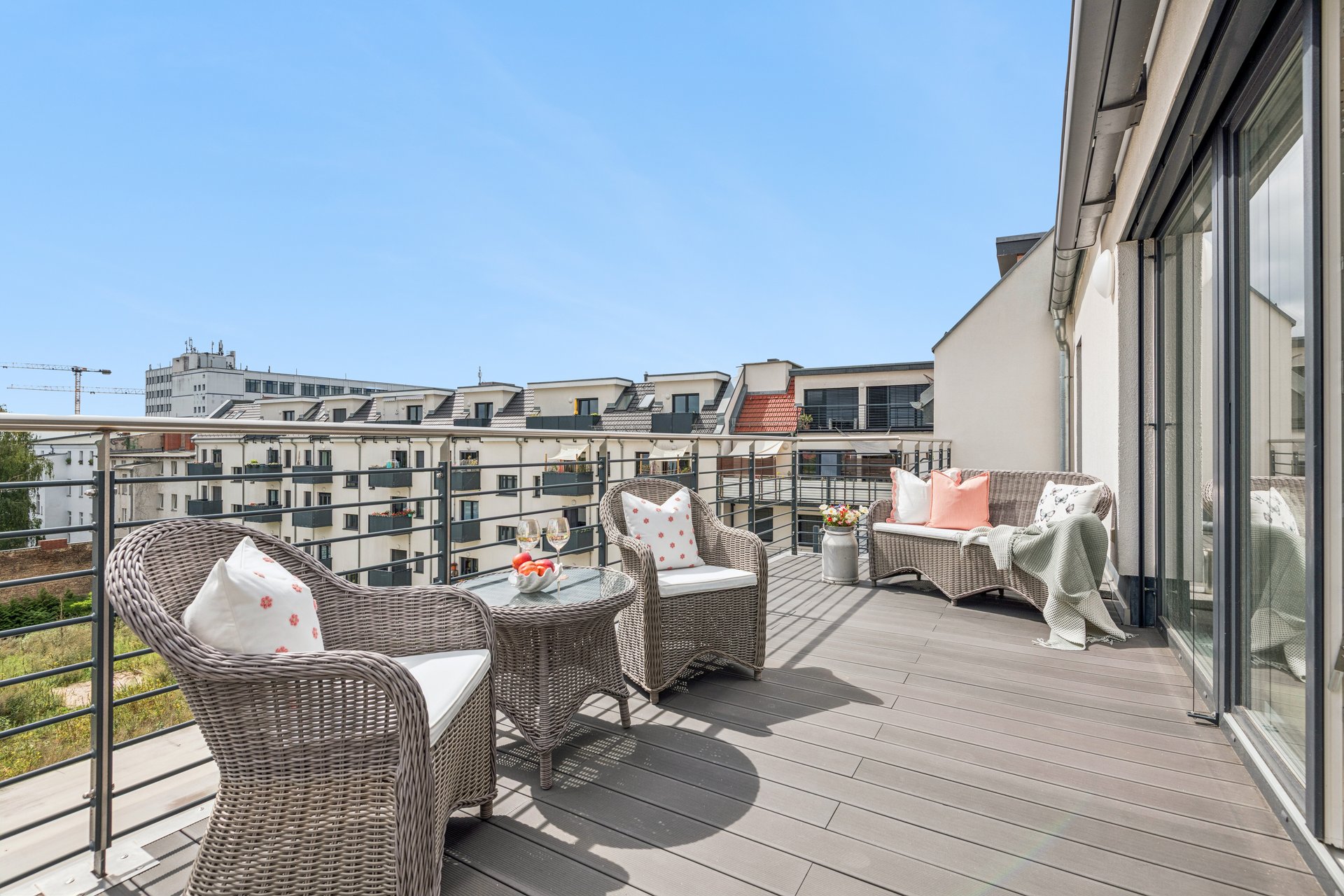 Sunny rooftop balcony with wicker furniture, a small table, and a view of neighboring apartment buildings under a blue sky. [AI-generated text]