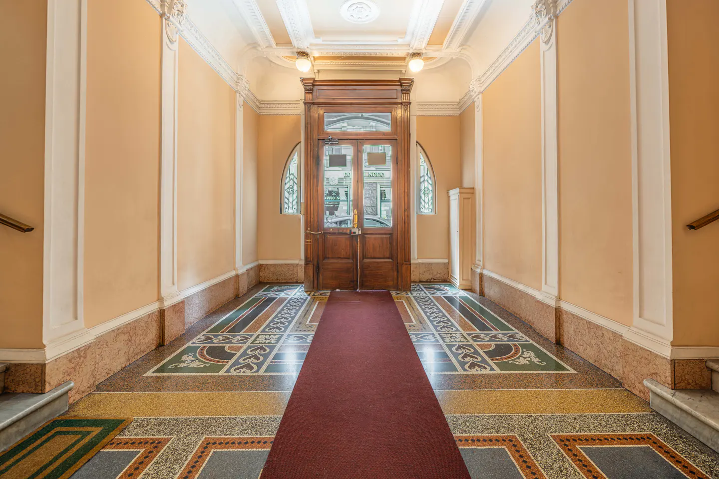 Hallway with peach walls, ornate trim, and a patterned tile floor. A red carpet leads to a wooden door with glass panels.