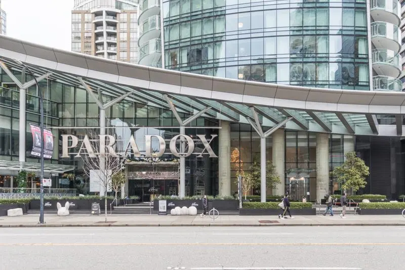 Exterior view of the Paradox Hotel with glass facade and silver signage. People walk on the sidewalk in front of the building.