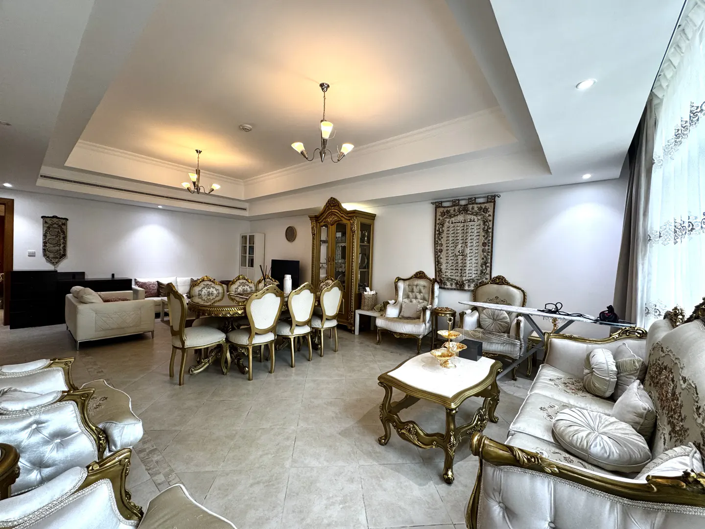 A spacious living and dining area with white and gold furniture, chandeliers, and tiled floors.