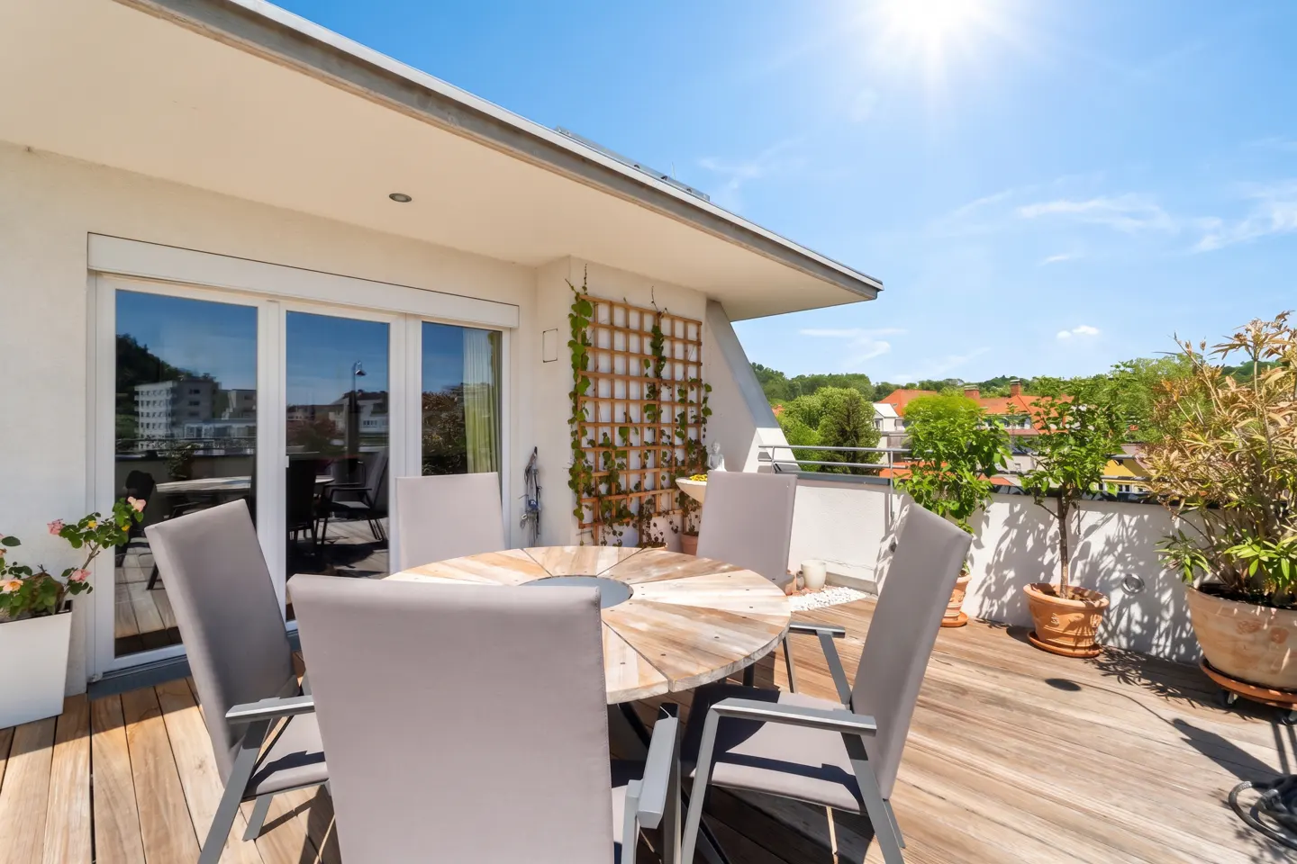 Sunny rooftop patio with a round wooden table, six gray chairs, potted plants, and a trellis with climbing vines.