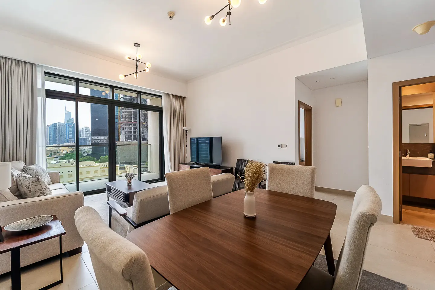 A bright living room with a wood dining table, beige sofa, and a large window overlooking a city skyline.