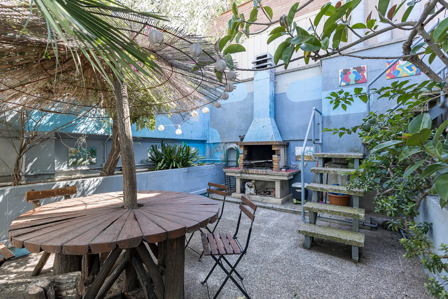 Outdoor patio with a round wooden table, chairs, and a brick barbecue against a blue wall. A tree grows through the table's center.