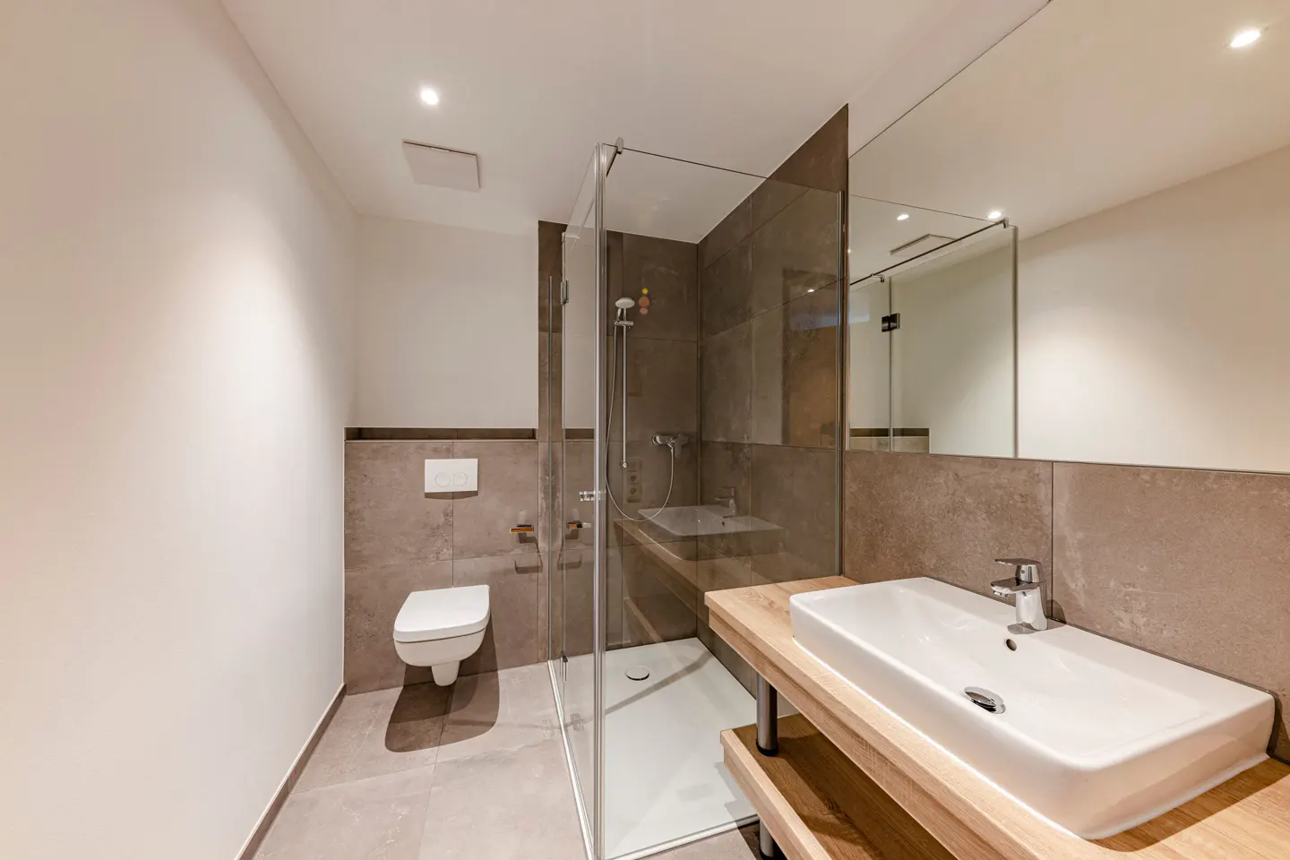 Bright bathroom with white walls, gray tile, and a glass-enclosed shower. A white sink sits on a light wood vanity. A toilet is visible to the left.
