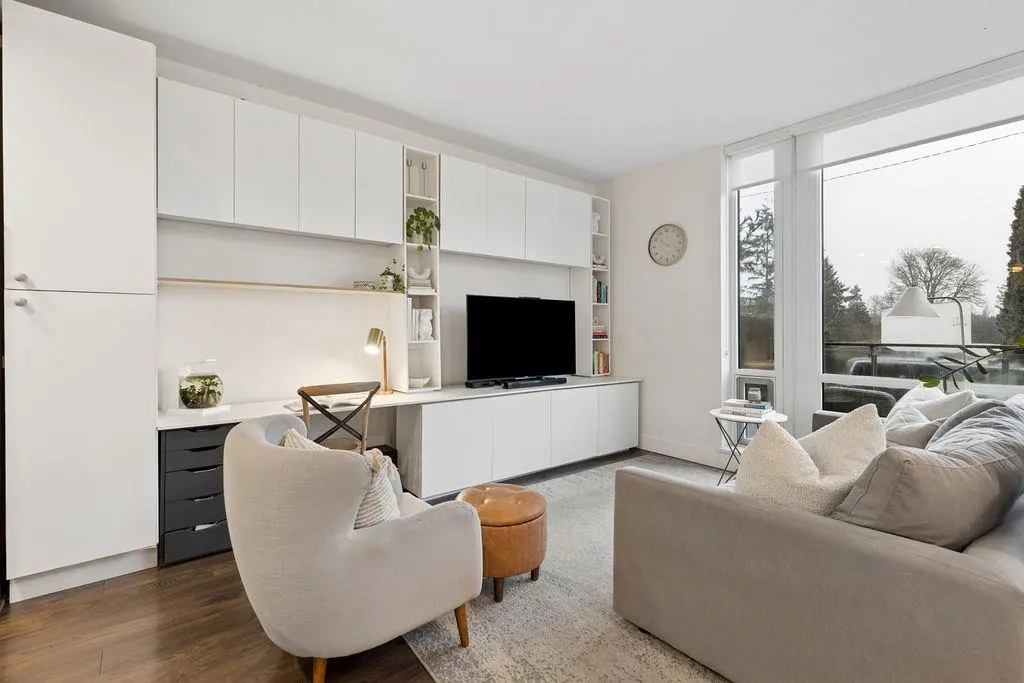 Living room with white built-in cabinets, desk, TV, gray sofa, chair, ottoman, and large window with outdoor view.