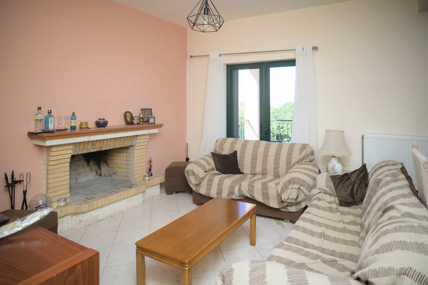 Living room with striped sofas, a wooden coffee table, and a brick fireplace with decor on the mantel. A window with white curtains is visible.