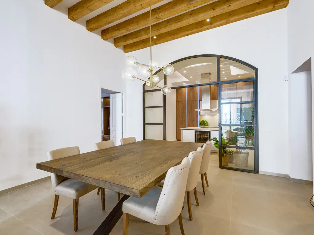 Bright dining room with a long wooden table, beige chairs, and a modern chandelier. Kitchen visible through a glass-paneled wall.