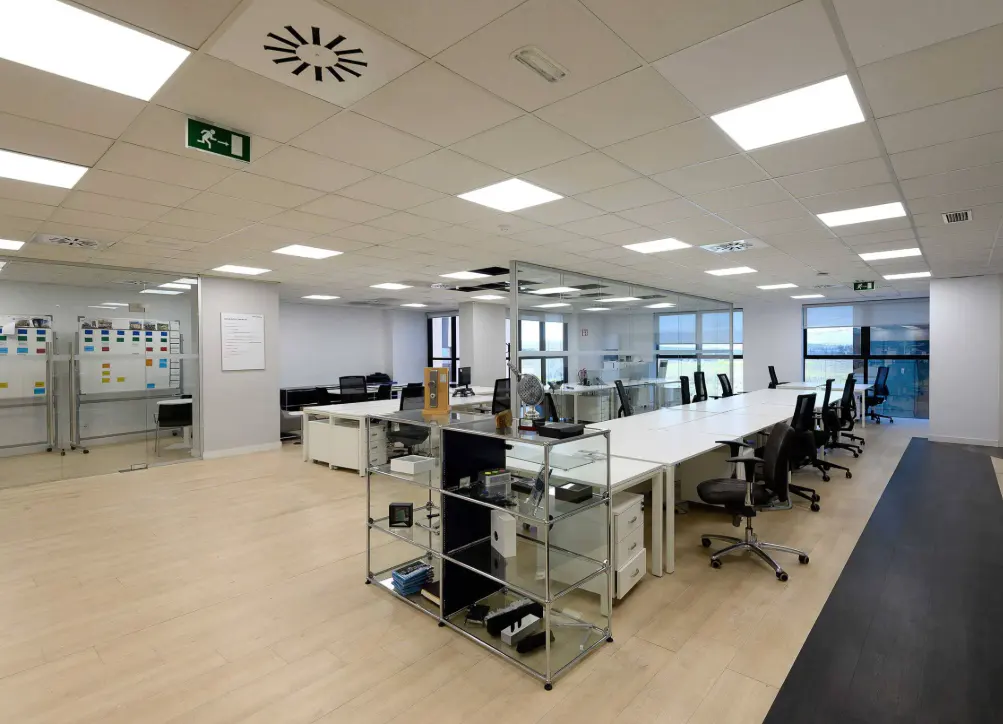 Bright, modern office space with white desks, black chairs, and light wood floors. A glass-walled conference room is visible in the background.