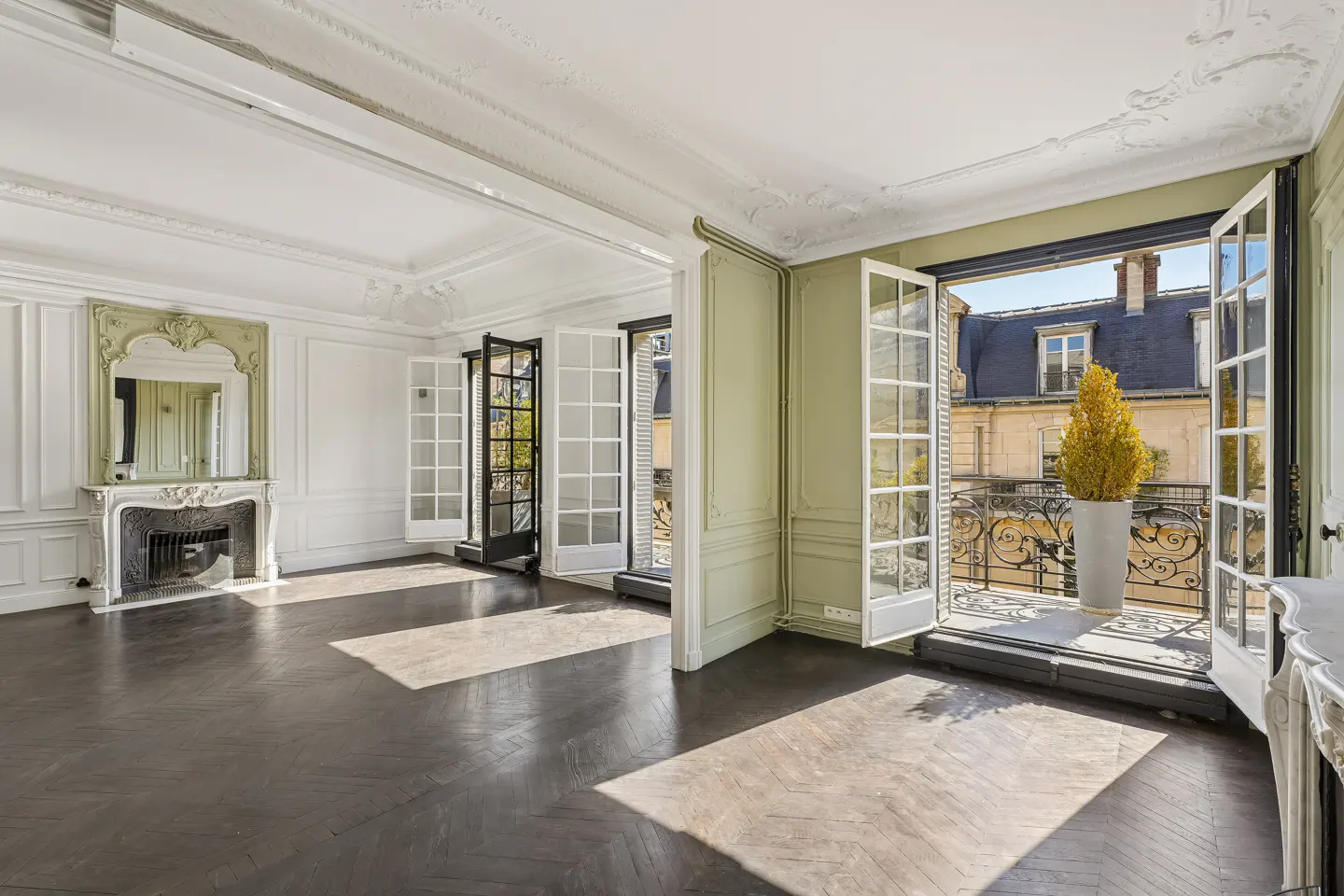 A bright, empty room with dark wood floors, white walls, and a fireplace. Open French doors lead to a balcony with a view of buildings.