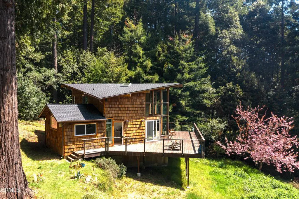 Exterior view of a wood-shingled house with a deck, surrounded by trees and greenery.