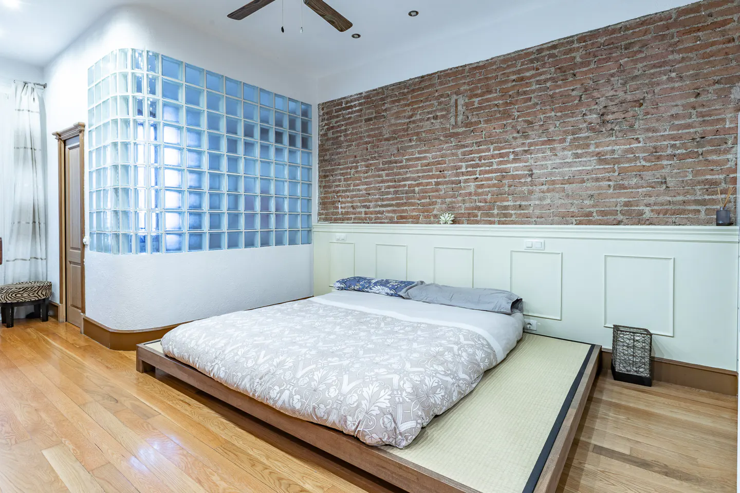 Bedroom with a low platform bed, brick wall, and glass block window. The floor is hardwood.