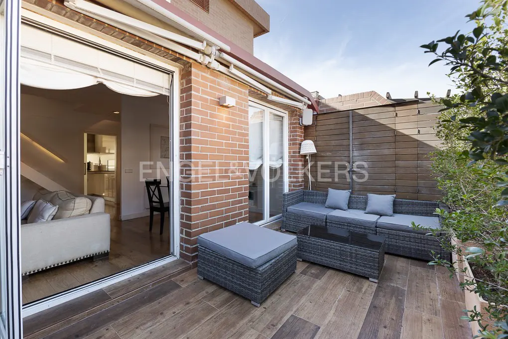 Outdoor patio with wicker furniture, wood flooring, and brick wall. Sliding glass doors lead to the interior. Green plants add a natural touch.