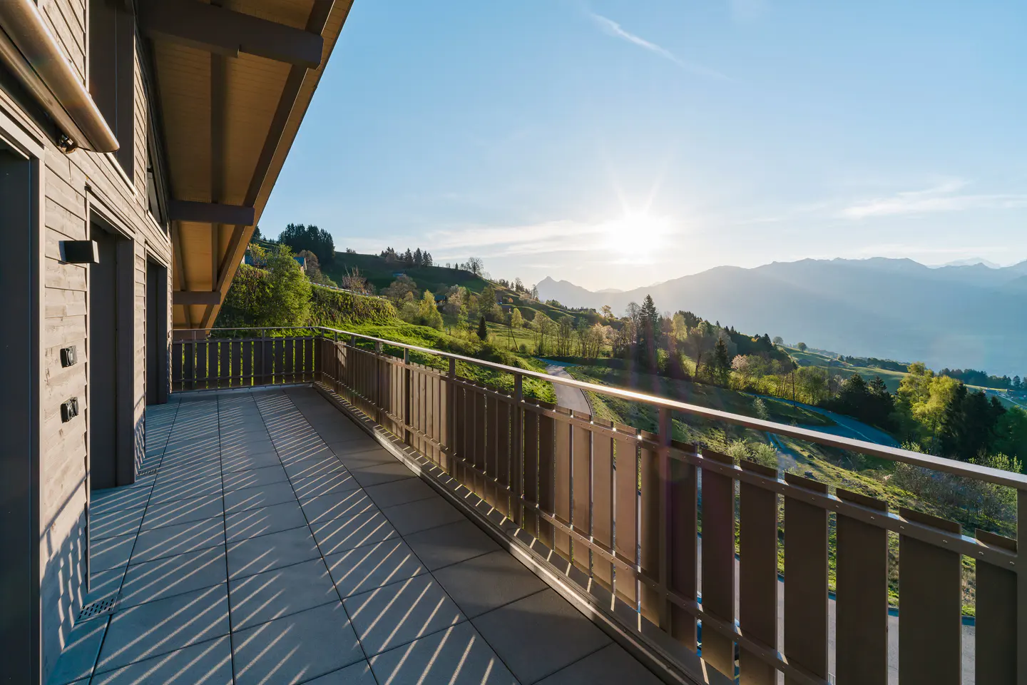 Balcony view of mountains and trees. Brown railing casts shadows on gray tiles. Blue sky with sun.