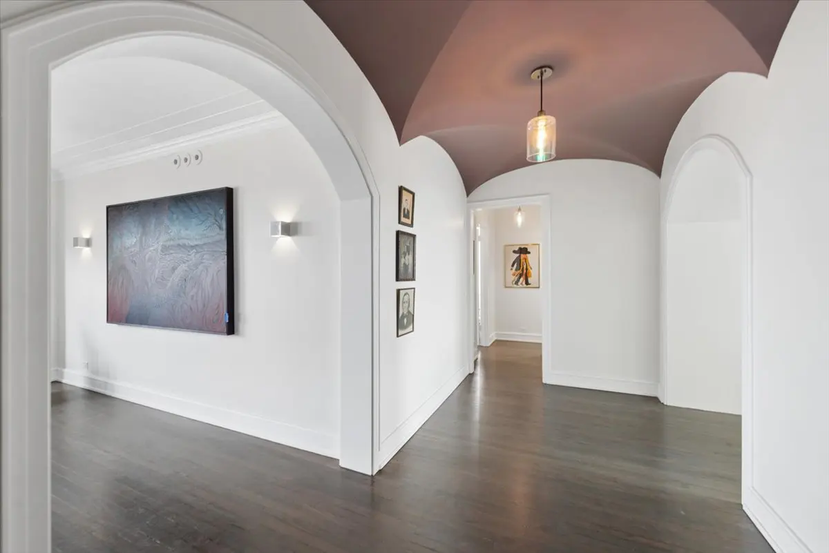 Hallway with white walls, dark wood floors, and arched doorways. Artwork hangs on the walls, and a pendant light illuminates the mauve ceiling.