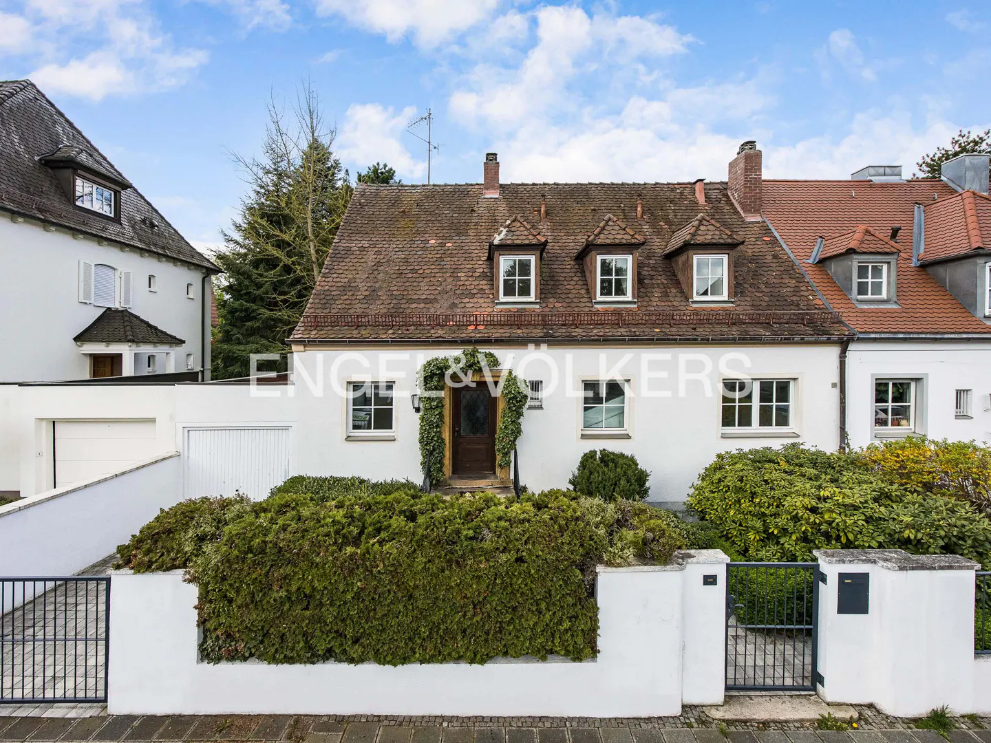 Exterior view of a two-story white house with a brown roof, dormer windows, and a green hedge in front. The Engel & Völkers logo is visible.