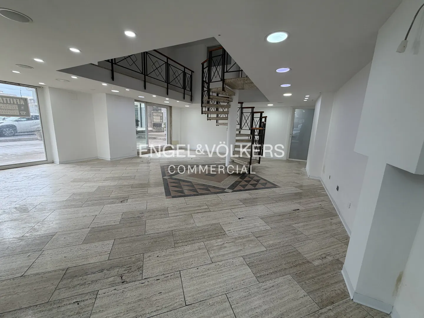 Bright commercial space with travertine floors, white walls, and a spiral staircase. Engel & Völkers logo is visible.