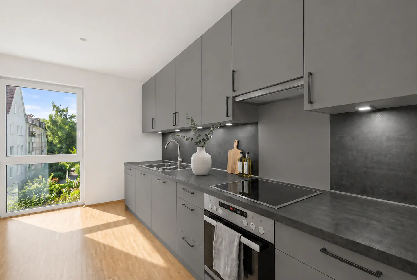 Modern kitchen with matte grey cabinets, dark countertops, built-in oven, and a large window overlooking greenery.