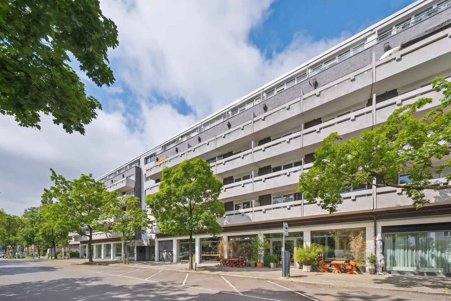 Exterior view of a long, gray apartment building with balconies and trees in front. Street-level shops have outdoor seating. Blue sky with clouds.