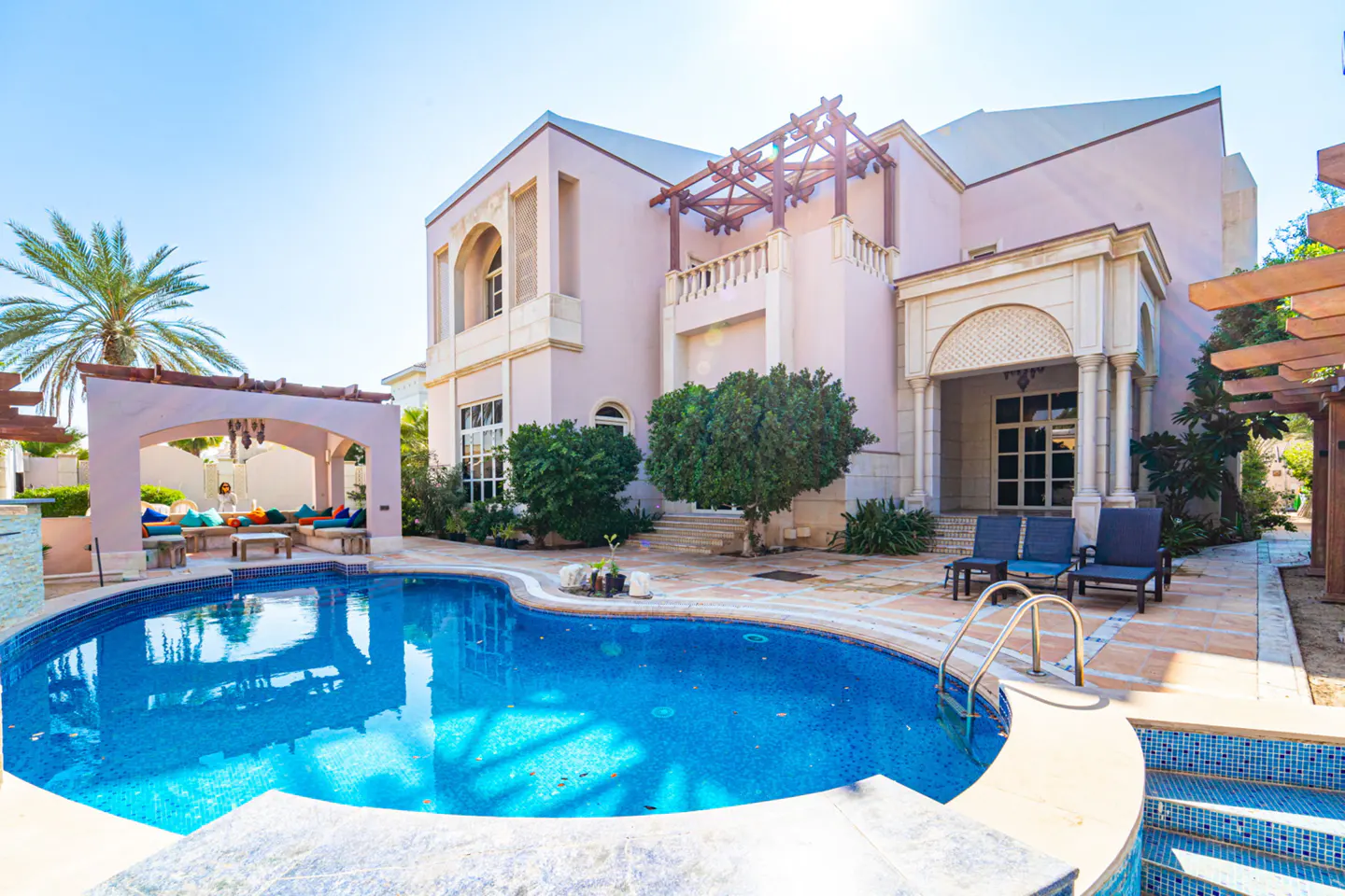 Exterior view of a two-story pink house with a blue tiled pool, lounge area, and palm tree under a clear blue sky.