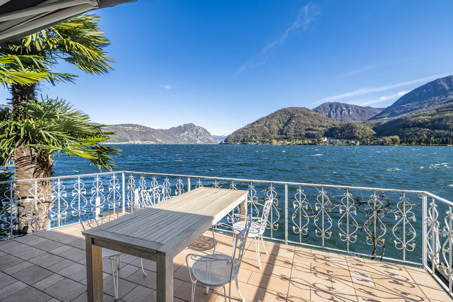 A sunny terrace with a wooden table and white chairs overlooking a blue lake and mountains under a clear sky.