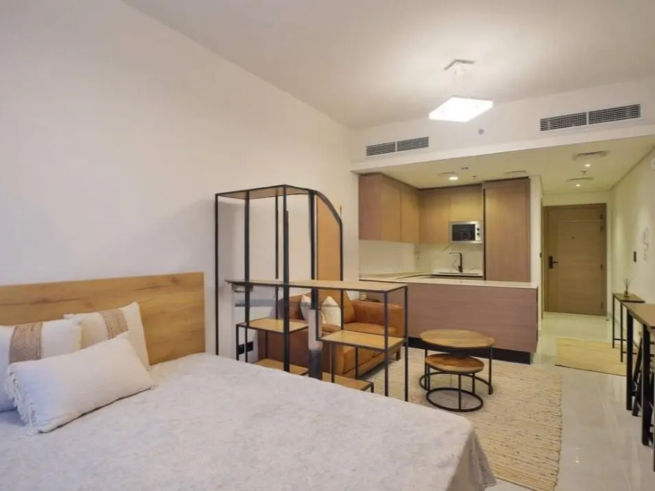 Studio apartment with a bed, sofa, shelving unit, and kitchen area. The walls are white and the floors are light-colored.