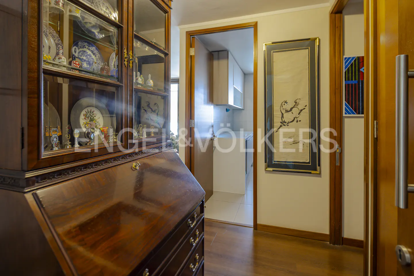 Interior view of a hallway with a wooden secretary desk, framed art, and a glimpse into a kitchen.