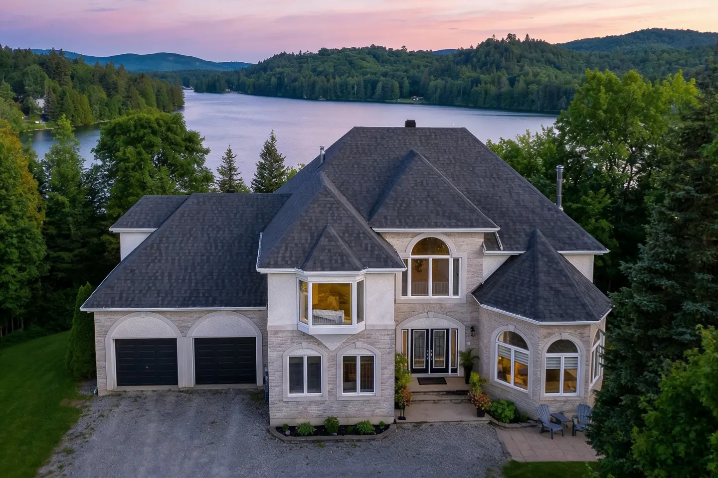Aerial view of a large, two-story stone house with a dark roof, surrounded by trees and a lake at sunset.