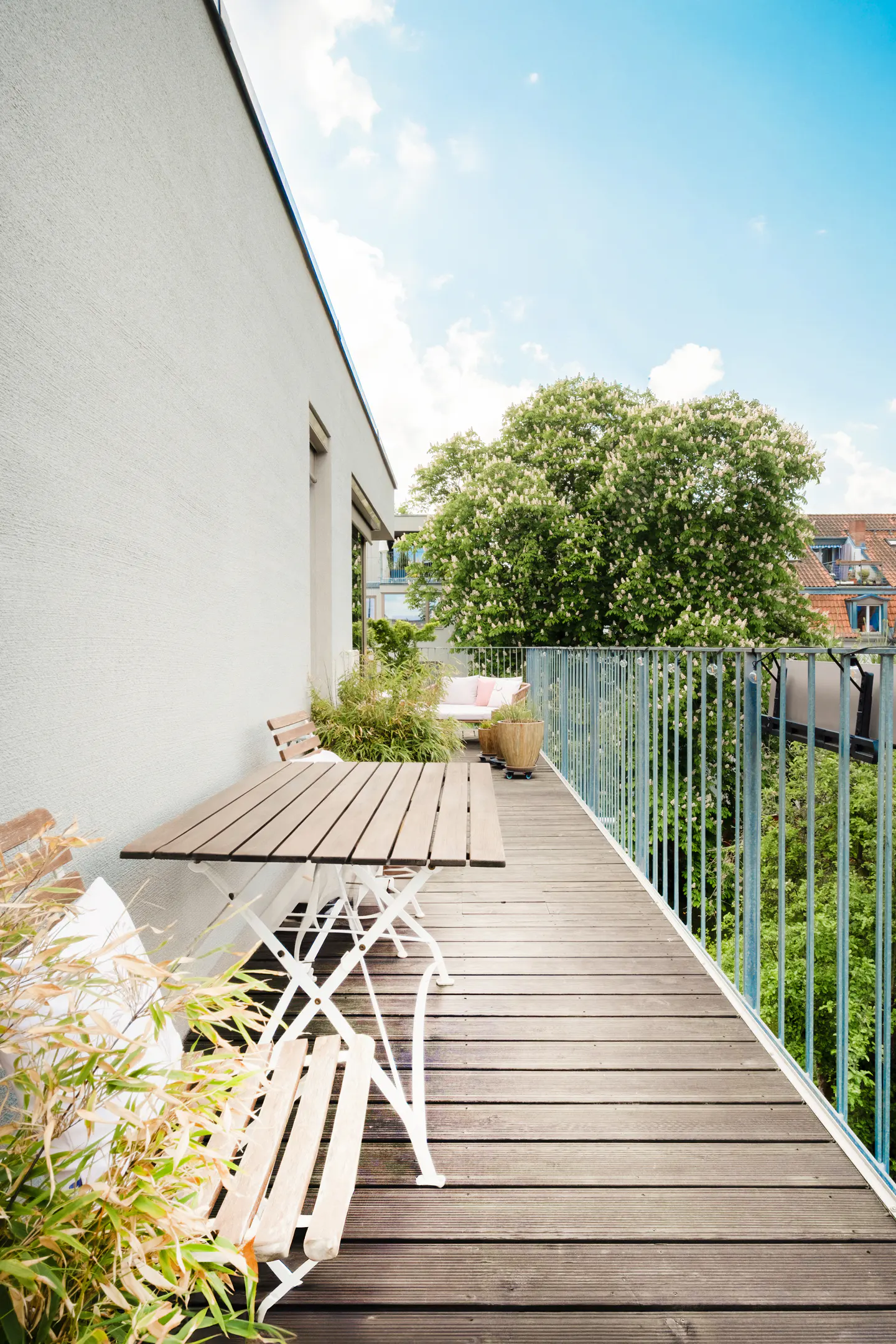 Spacious wooden balcony with a white table and chairs, overlooking lush green trees under a bright blue sky.