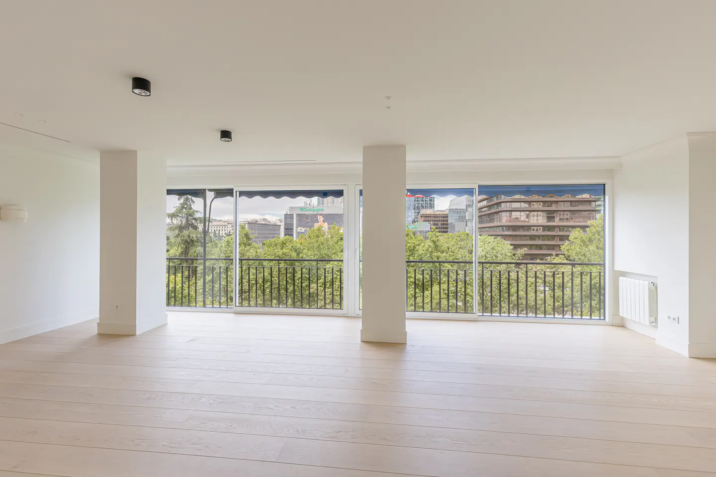 Bright, empty room with light wood floors, white walls, and large windows. A black railing overlooks green trees and city buildings.