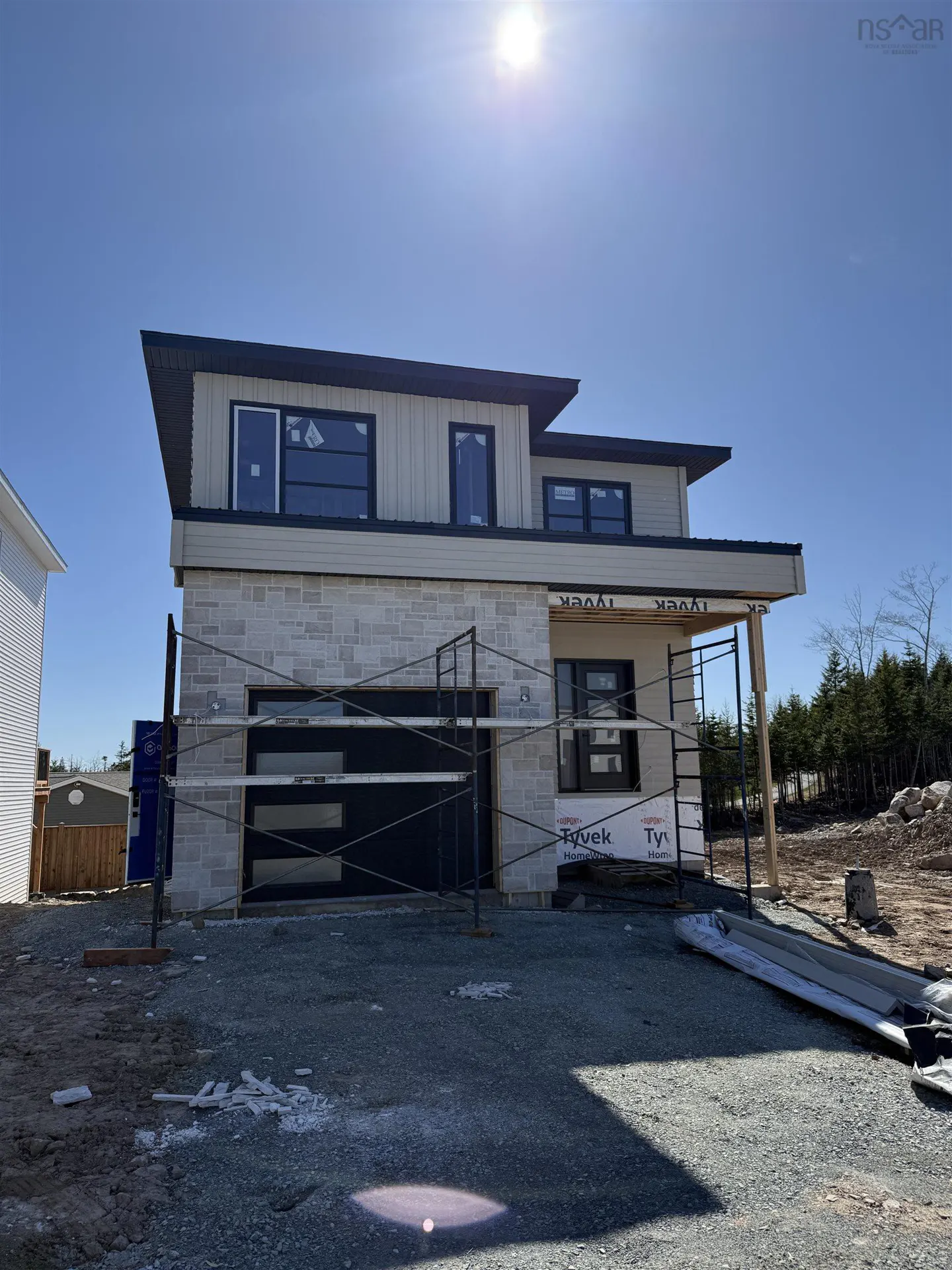 Two-story house under construction with stone and beige siding, black trim, and a black garage door on a gravel lot.