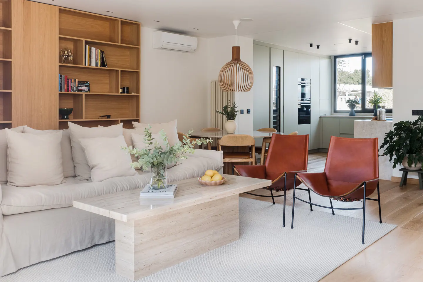 Bright, open-concept living space with a beige sofa, travertine coffee table, and two brown leather chairs. A light wood bookshelf and kitchen are in the background.